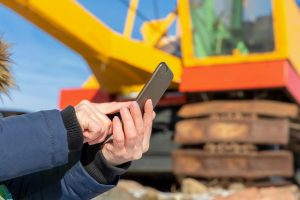 Person using smartphone near mini excavator at construction site.