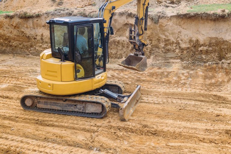 Small yellow excavator leveling soil on construction site.