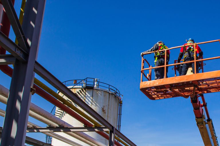 Industrial workers inspecting equipment on a lift at a construction site under a clear blue sky.
