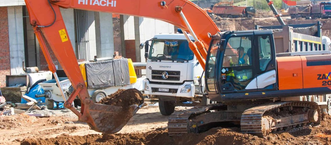 Orange Hitachi excavator digging at a construction site with trucks and equipment nearby.